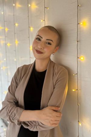Valerie with her head shaved standing against a wall with string lights