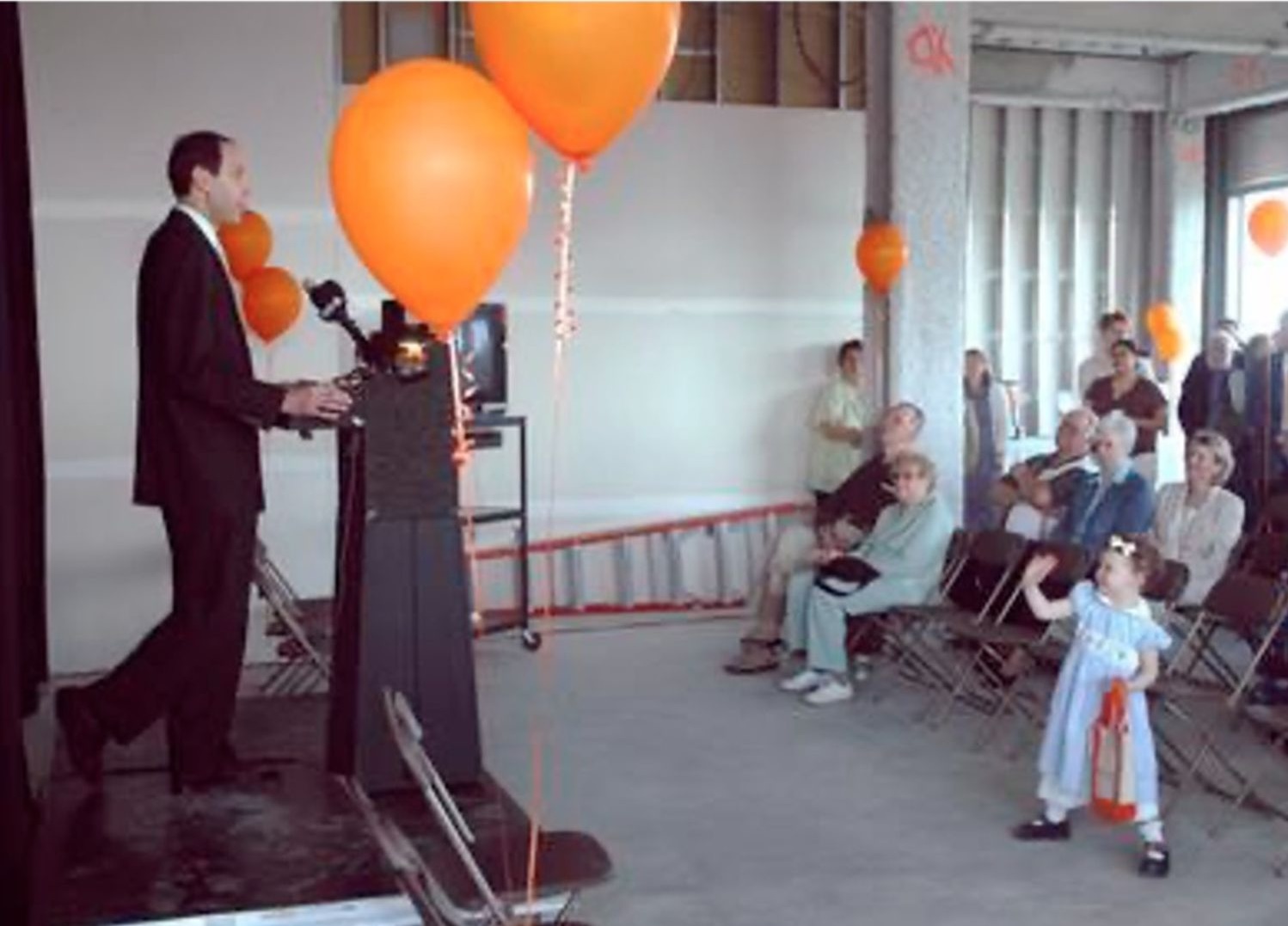 A man stands at a speaking podium, surrounded by orange balloons. He's facing a small crowd of people sitting in folding chairs or standing as they listen to him. A young girl wearing a dress and hair bow waves in the foreground.