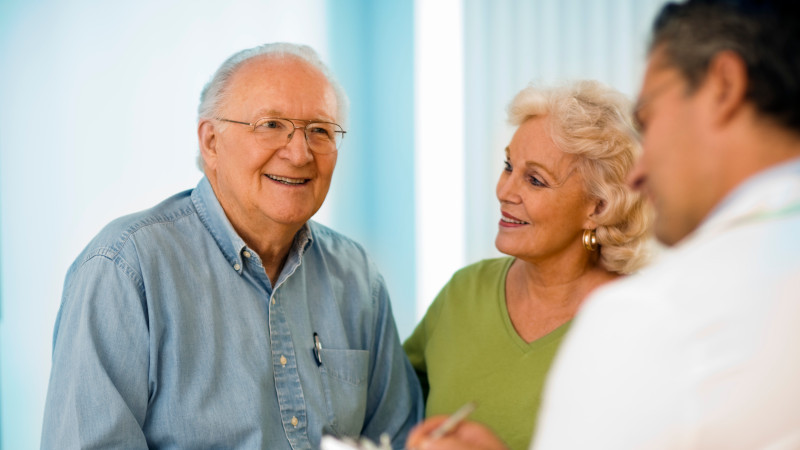 An older, white man smiles and is joined by his wife, who is also white, as he speaks to his doctor