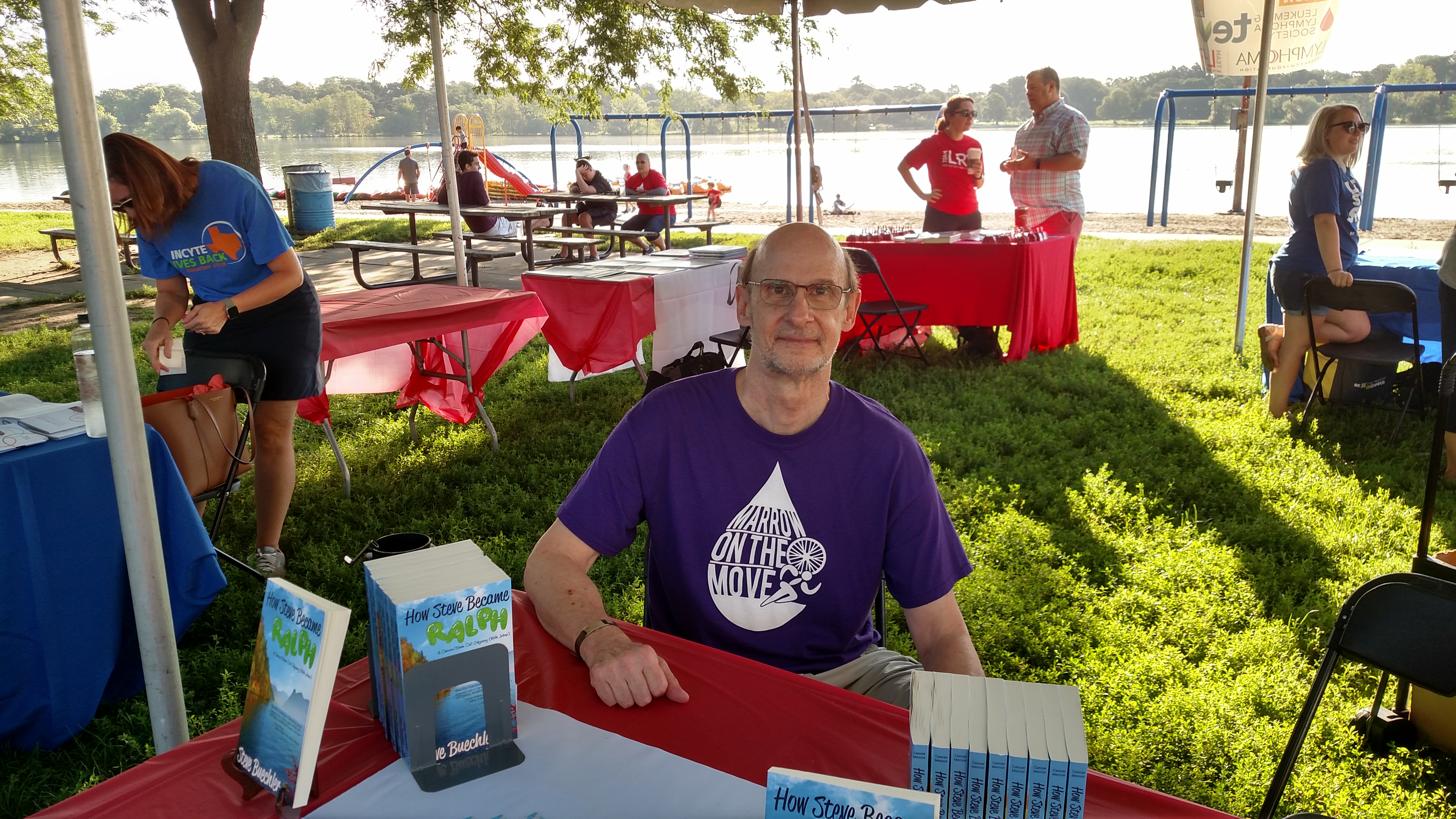 Steve, a white man with glasses, sits at a table with a stack of books. The table is surrounded by several other tables in a park. 