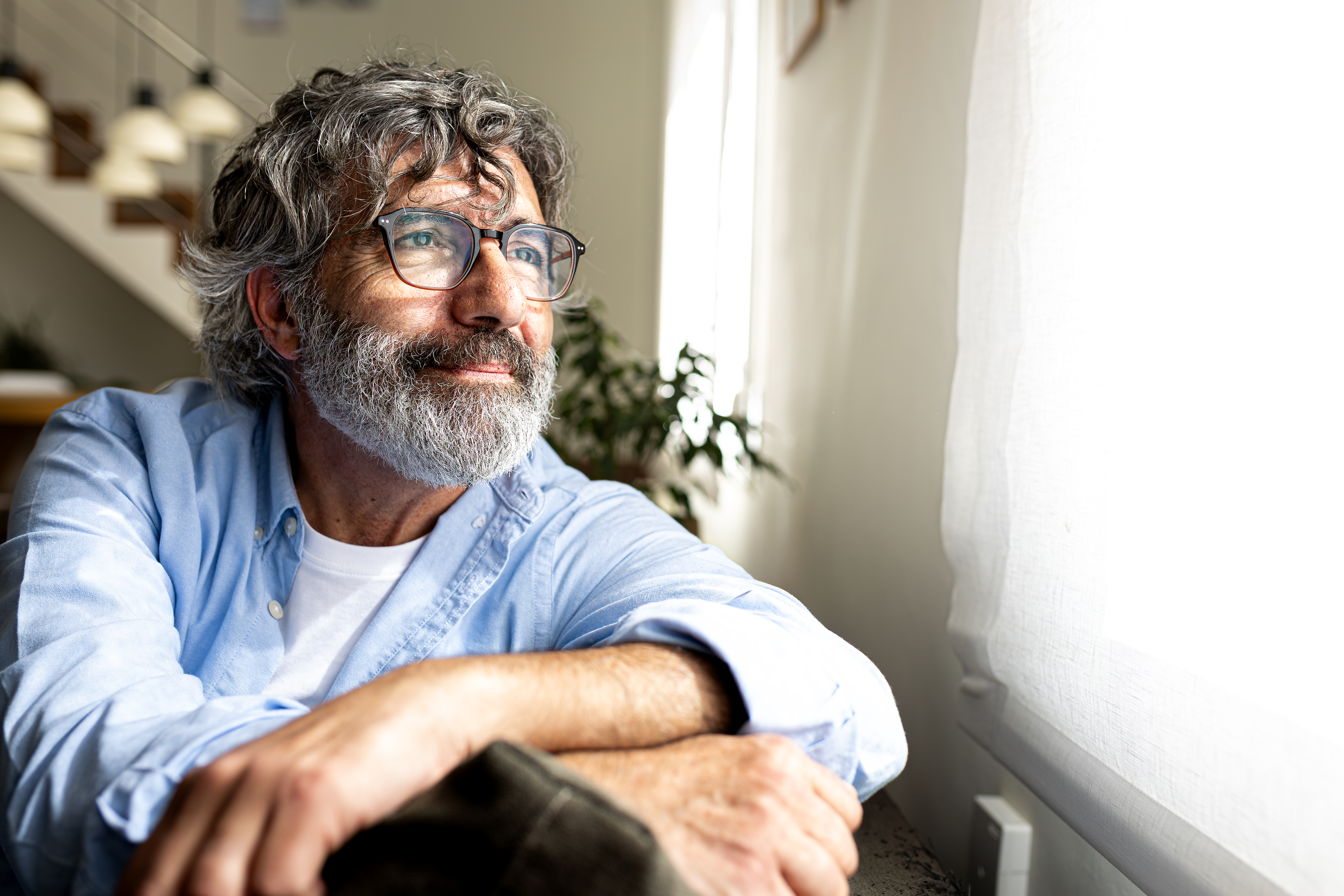 A content-looking man with long gray hair and a thick beard sits at home and looks out the window