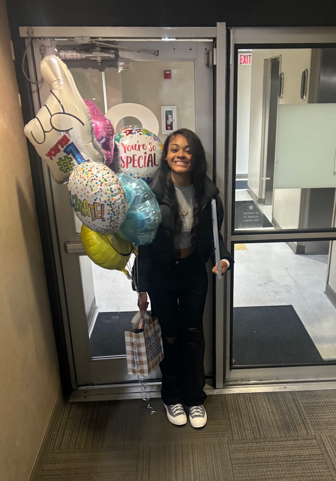Smiling black teen girl with lymphoma in black jacket and pants holding mylar balloons and a gift bag