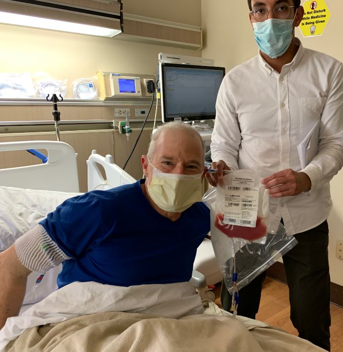 Man in blue shirt and mask in a hospital bed with a medical worker holding a bag of stem cells
