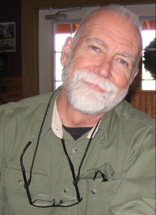 older white gentleman with white beard and mustache wearing a long-sleeved green shirt with a pair of glasses hanging around his neck