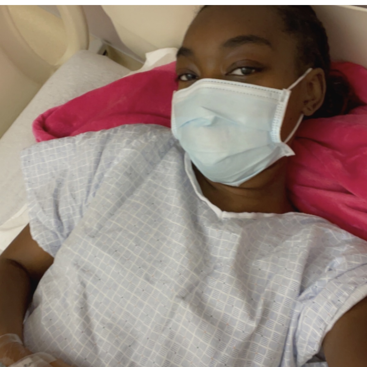 young black woman wearing a mask and hospital gown lying on a bed with a pink blanket