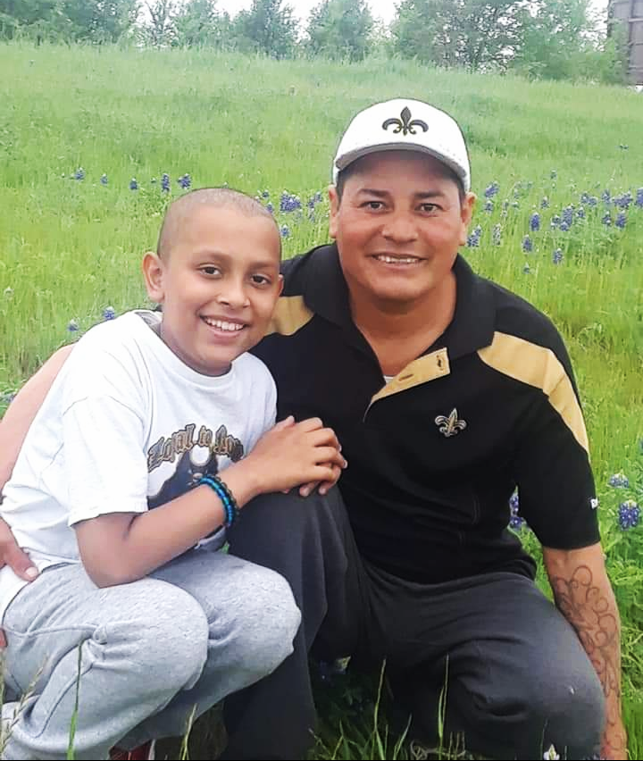 young hispanic child with little hair in white t-shirt and sweat pants with his father who is wearing a New Orleans Saints hat and a black and gold shirt, both in a field of bluebonnets