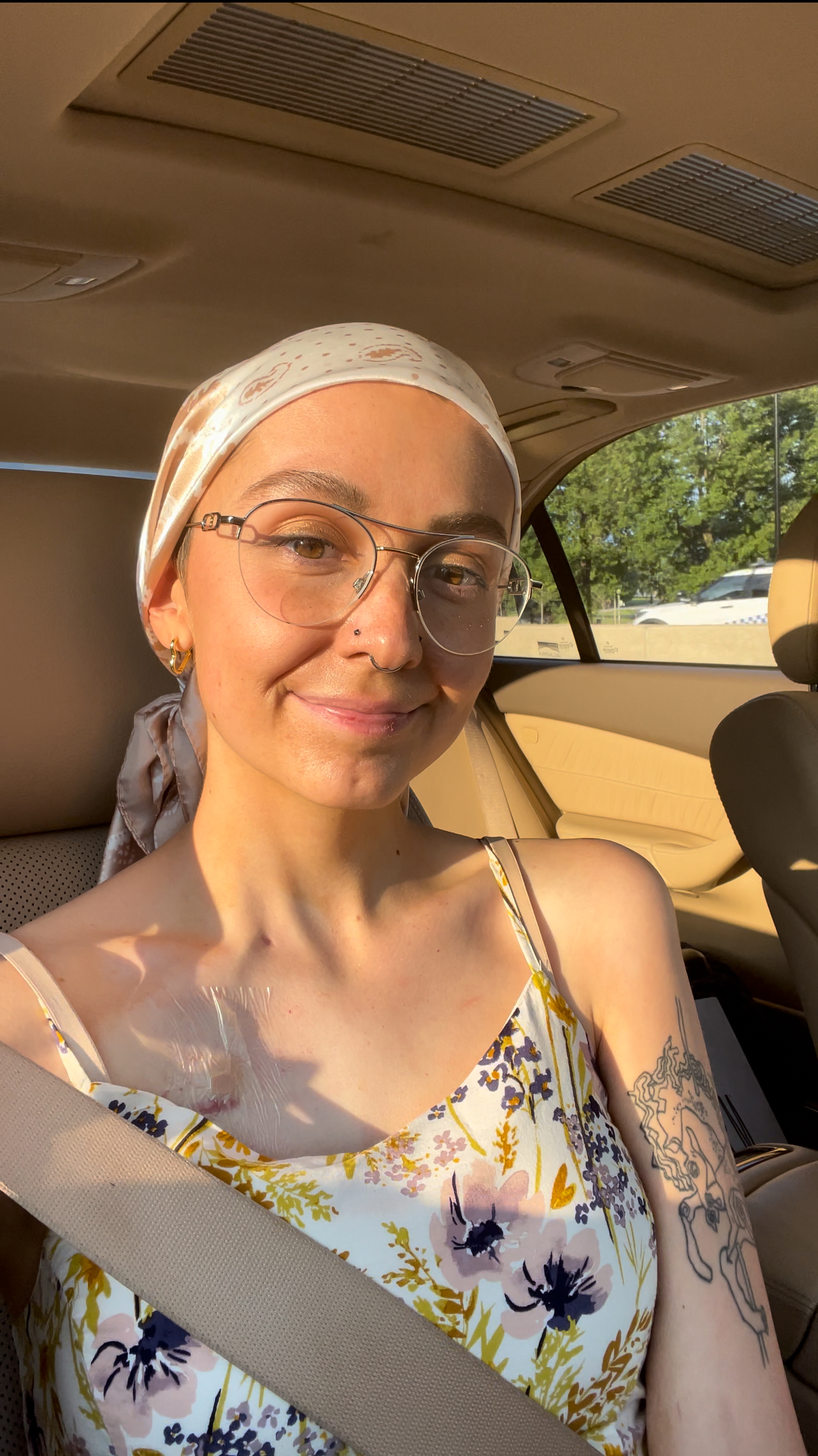 young white woman sitting in a car wearing a head scarf, wire rim glasses, a floral sundress witha nose ring