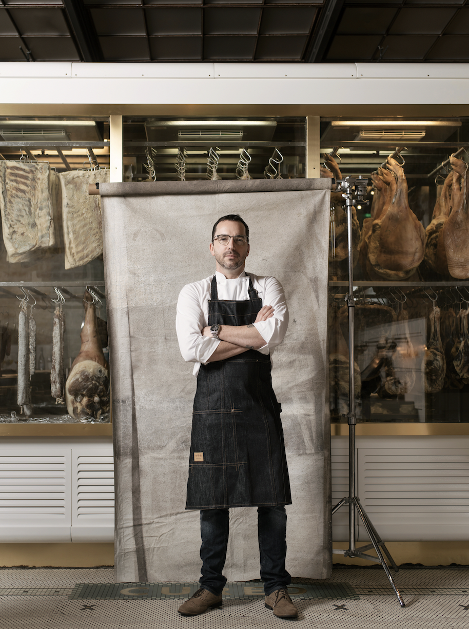 Steve young white man with short hair glasses and a beard and mustache wearing a white chef's coat under a denim apron standing in front of a canvas that is front of windows showing meats being cured