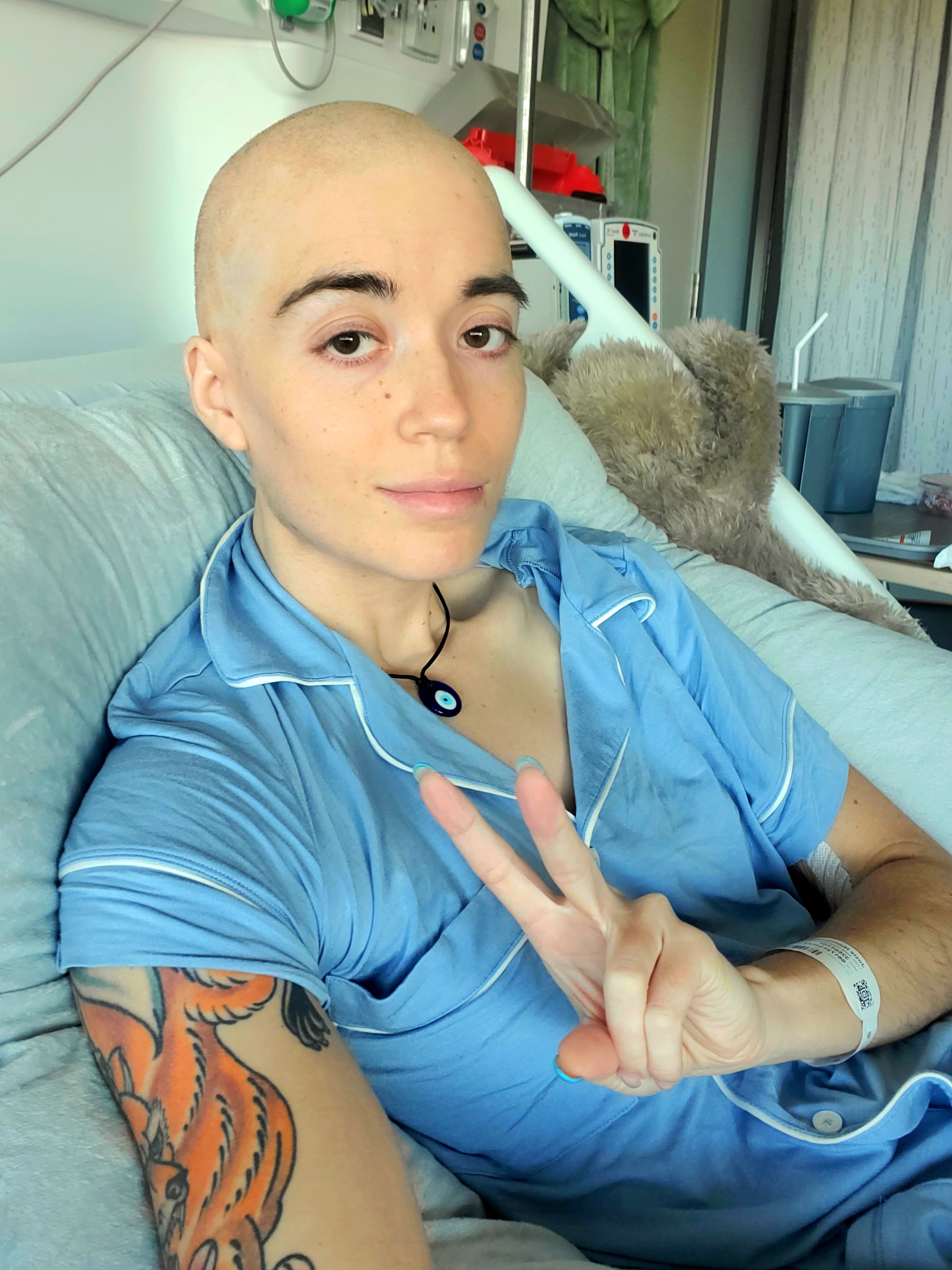 Josie young white woman bald with an orange tatoo and wearing blue pajamas in a hospital bed giving the peace sign