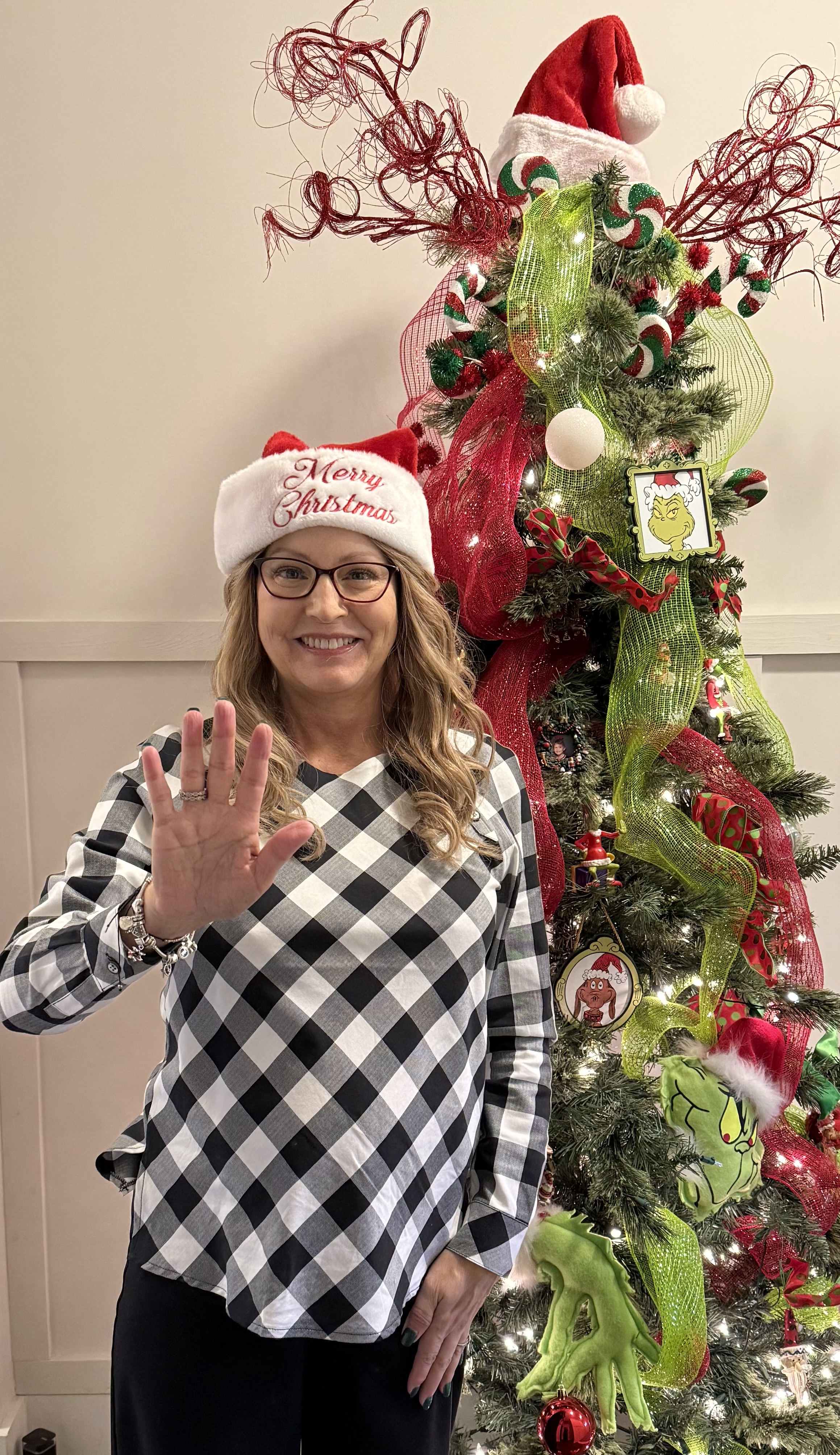 Lori middle aged white woman with long blond hair wearing a Merry Christmas santa hat and glasses and black and white geometric blouse standing in front of a Christmas tree