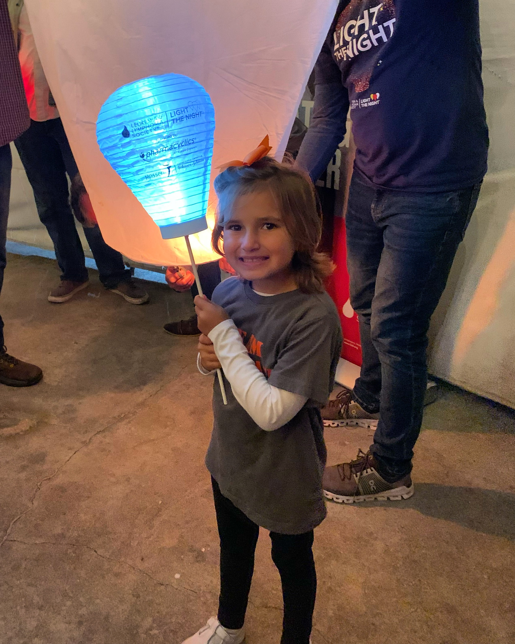 young smiling white girl in a gray shirt over a white long sleeved shirt with and orange bow in her hair and holding a LTN balloon