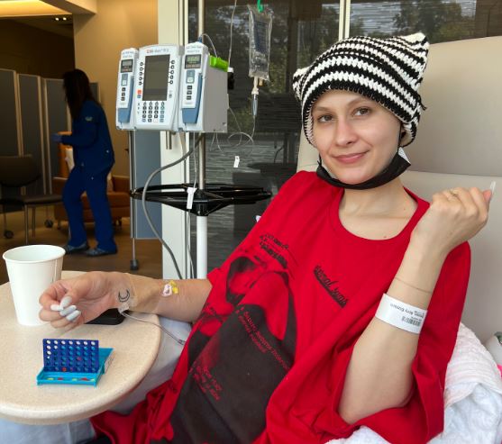 young white woman wearing a black and white knit cap and a red t-shirt and black short with painted white fingernails laying in an infusion chair