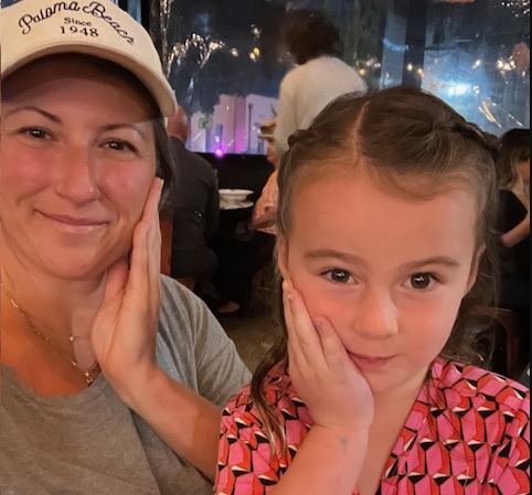 young white woman wearing ball cap and greenish tshirt with daughter in a pink and black top and both have a hand up against their cheeks