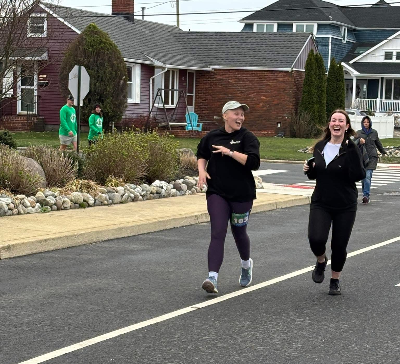 Ally two young white women running along a street one bald and wearing a ball cap both in dark running gear