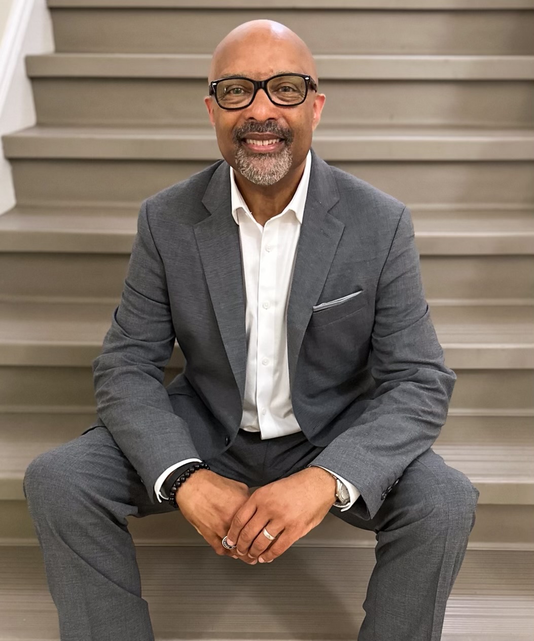 Arturo Pierre middle aged black man bald with mustache and beard wearing black glasses and a grey suite and white shirt sitting on steps