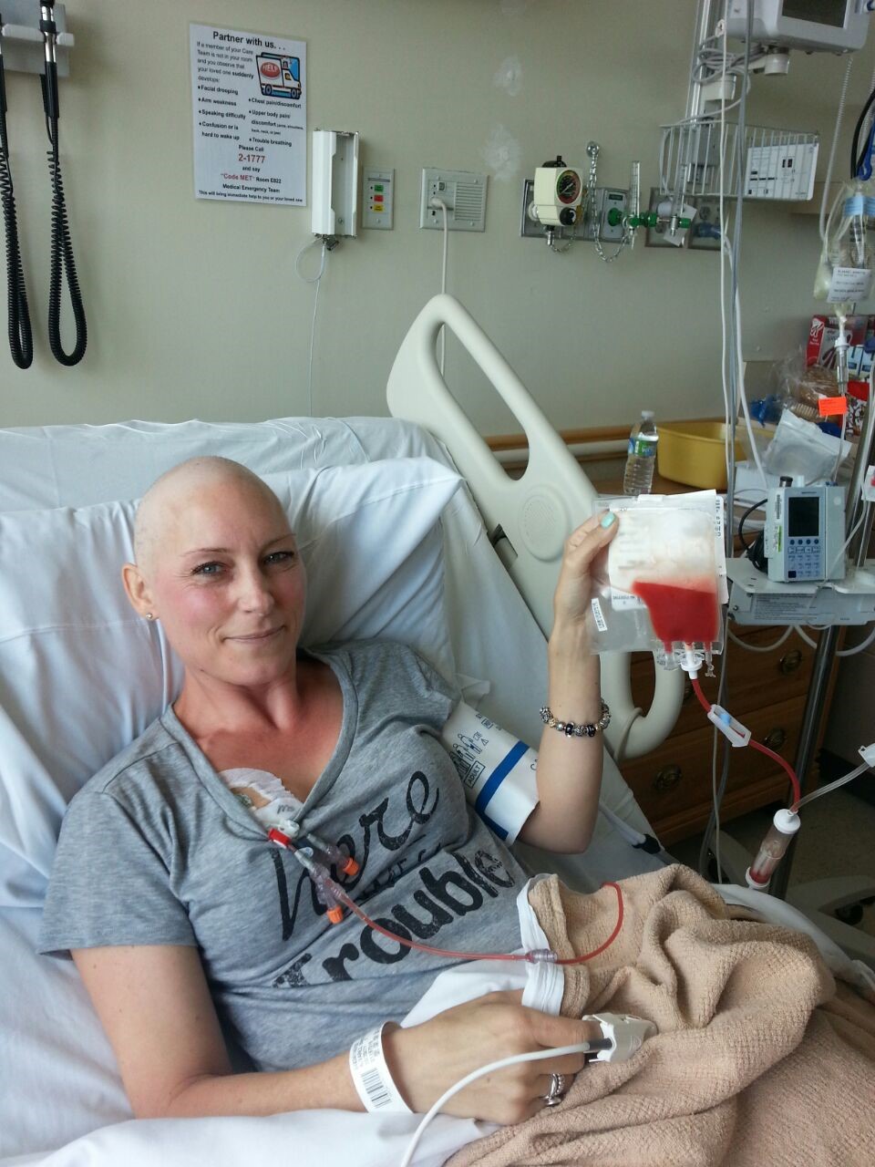 Babette young white woman who is bald waring a grey t-shirt and lying in a hospital bed holding an infusion bag