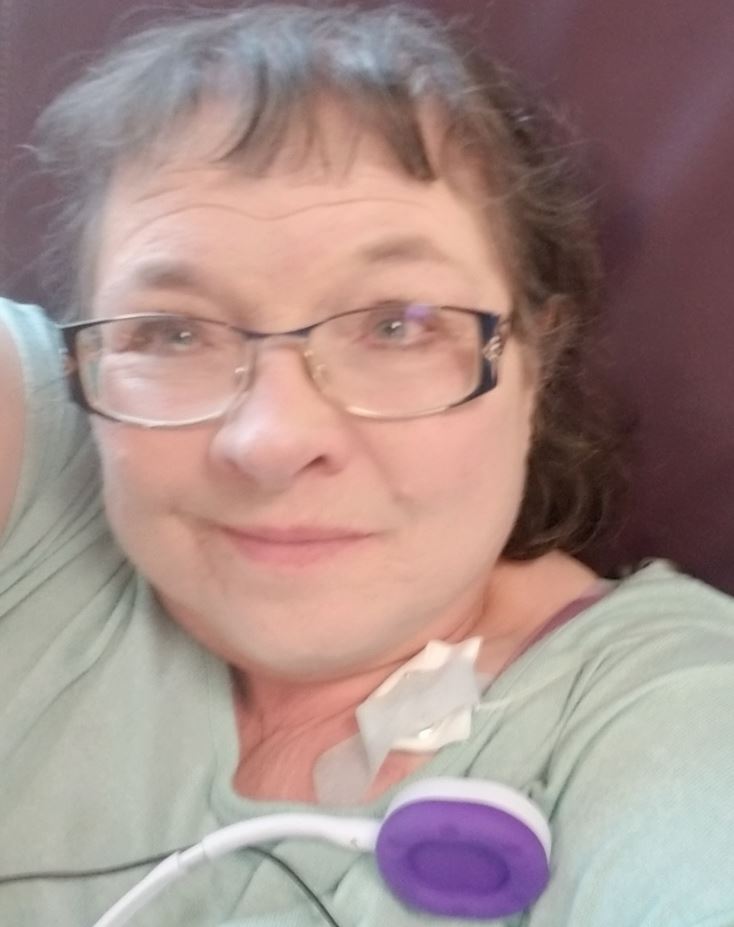 Dixie older white woman with brown hair and glasses wearing a green shirt laying against a brown pillow with an earbud laying on her chest