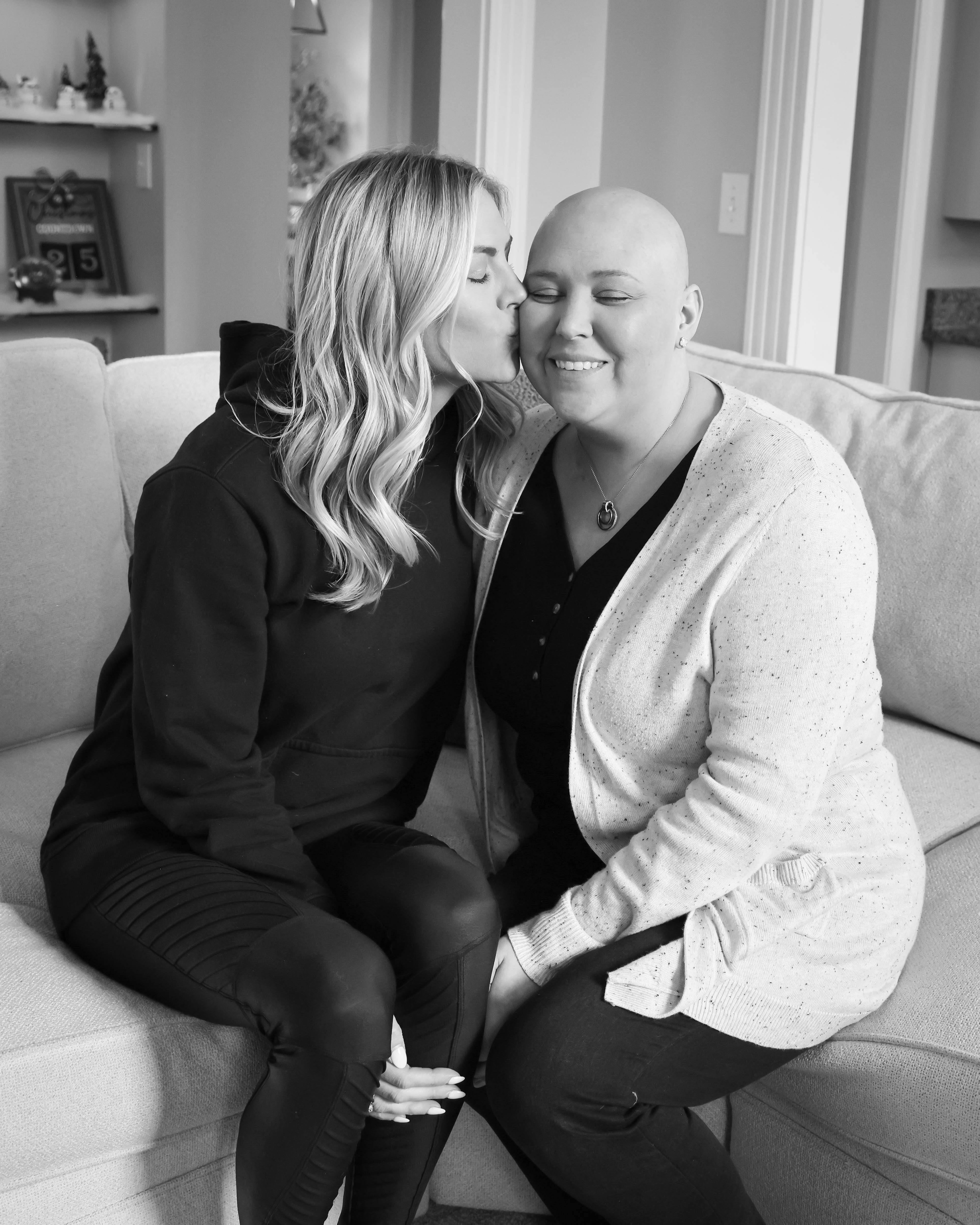 Janie young white woman who is bald wearing a black shirt and a white speckled cardigan being kissed by a young white woman with long blond hair wearing a black hoodie both sitting on a couch