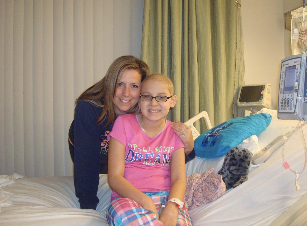 Kendra woman and young hispanic girl on a hospital bed girl has glasses and wearing a pink shirt and plaid pajama bottoms with turqouise pillow on bed