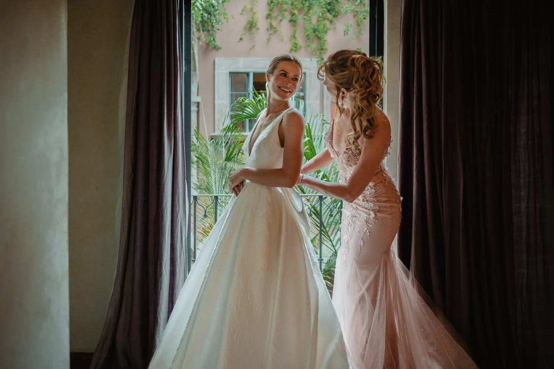 Laura young white woman in a white wedding dressing looking at her mother who has a curly hairdo and wearing a pink formal dress both standing in front of a balcony