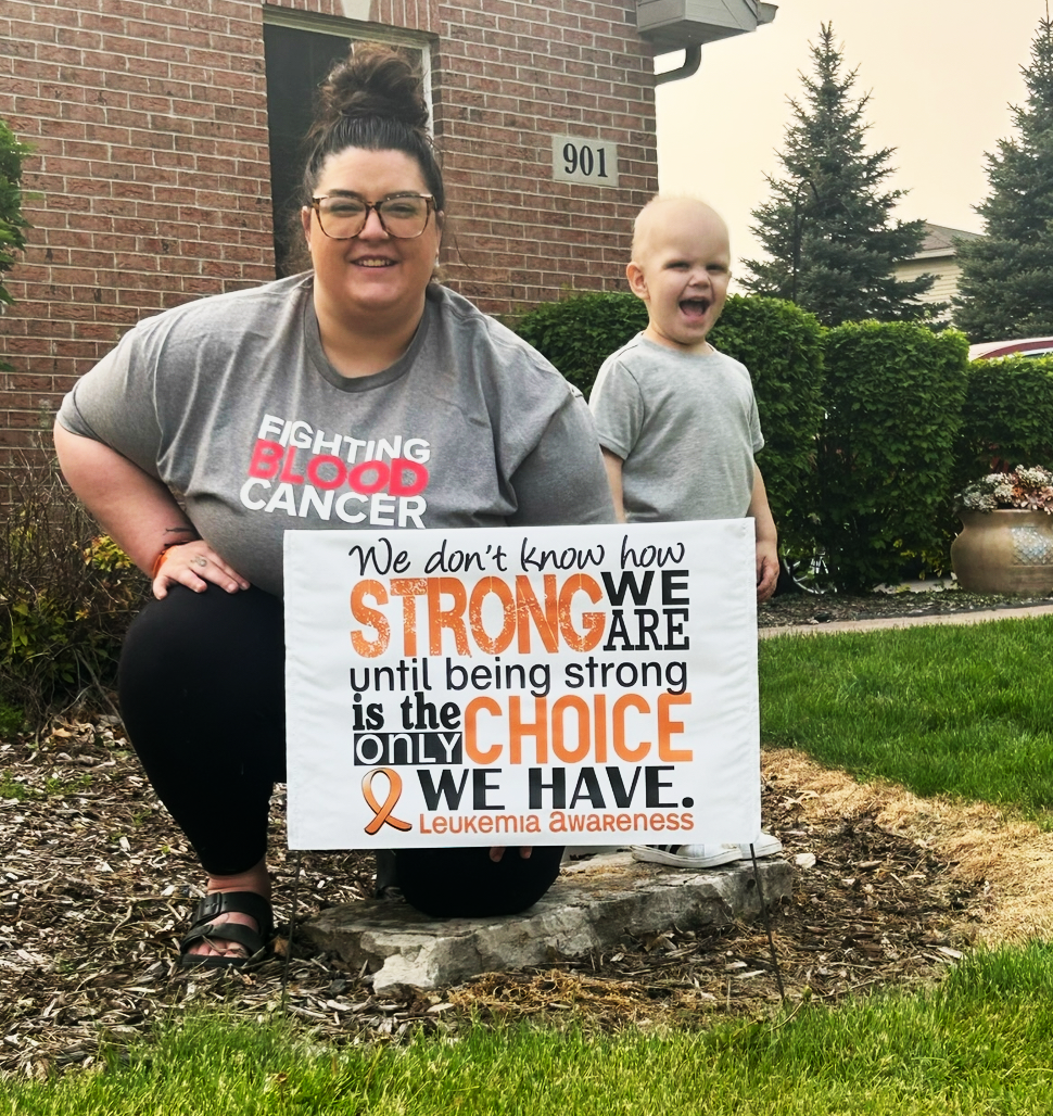 Maverick young white mother with a bun and glasses wearing gray blood cancer shirt holding a leukemia sign with her young son next to her in a gray shirt and smiling