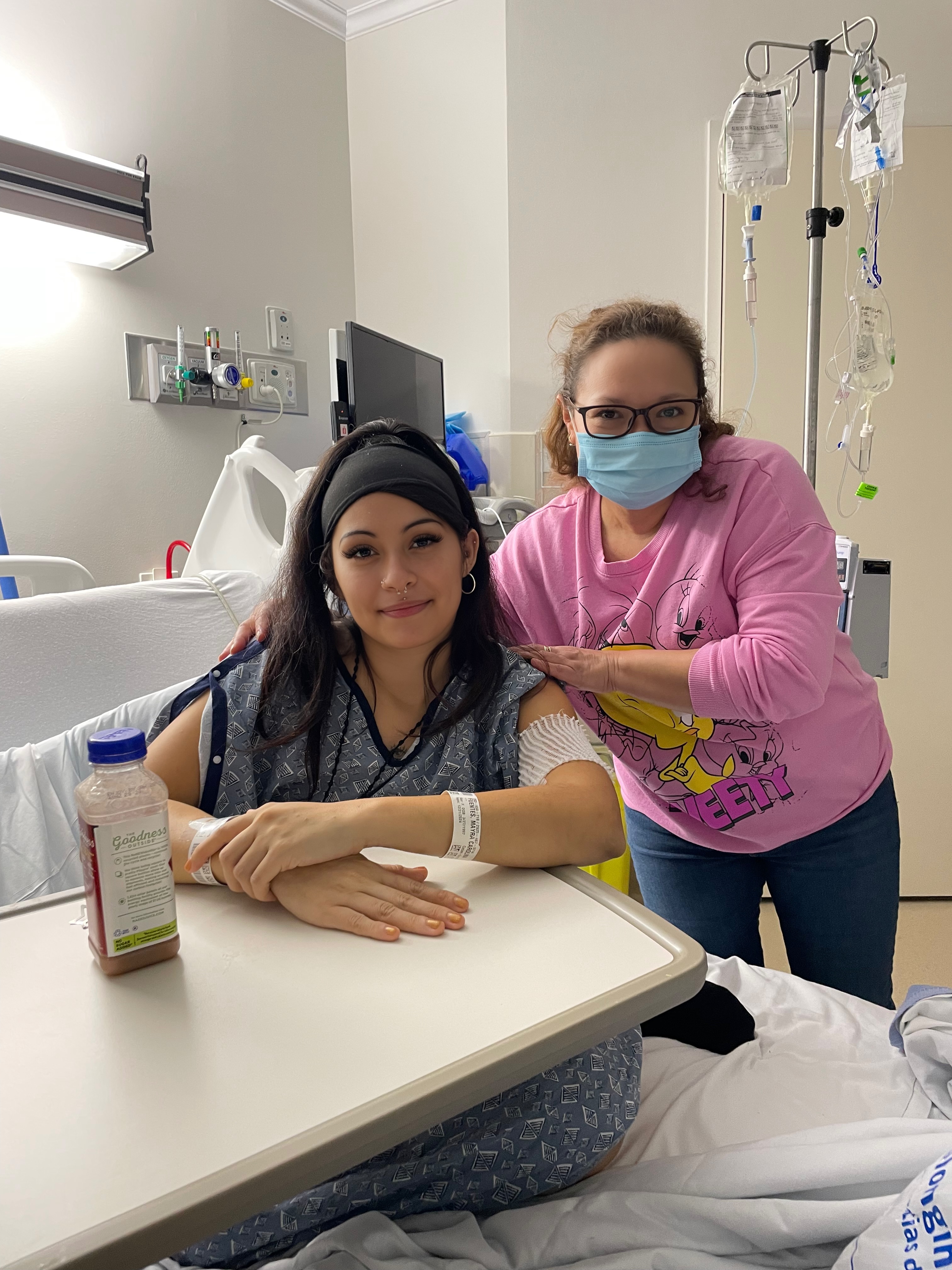 Mayra young hispanci woman sitting in a hospital bed wearing a black headbank and blue hospital gown and has three face piercings with another woman with face mask and pink shirt next to her