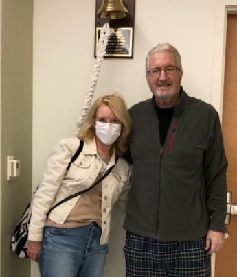Rex older white couple standing in a hospital room man has white hair and glasses wearing a gray jacket and pajama pants woman has face mask