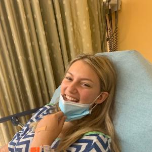 Blonde teen girl with leukemia in blue and white medical gown pulling down a face mask