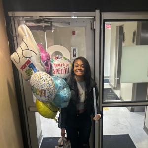 Smiling black teen girl with lymphoma in black jacket and pants holding mylar balloons and a gift bag
