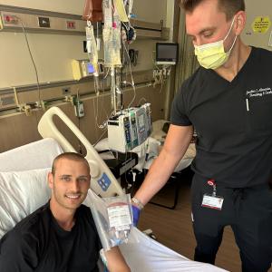 Young man in black t-shirt with leukemia in a hospital bed with a medical professional standing next to him wearing a mask