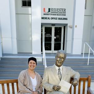 bald white woman wearing a knit cap tan sweater and white shirt sitting on a bench next to a statue