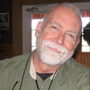 older white gentleman with white beard and mustache wearing a long-sleeved green shirt with a pair of glasses hanging around his neck