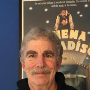 older white man with bushy eyebrows and a mustache wearing a black hoodie that says Italia standing in front of a movie poster