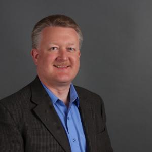 smiling middle aged white man with graying hair and mustache wearing a brown jacket and blue shirt
