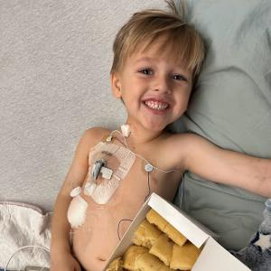 smiling young white boy laying in bed with a port in with a box of rolls