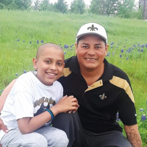 young hispanic child with little hair in white t-shirt and sweat pants with his father who is wearing a New Orleans Saints hat and a black and gold shirt, both in a field of bluebonnets
