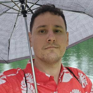 young man with scruffy beard and mustache in an orange and white shirt holding an umbrella in front of water