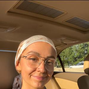 young white woman sitting in a car wearing a head scarf, wire rim glasses, a floral sundress witha nose ring