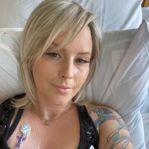 young woman with blond hair and long eyelashes lying in hospital bed wearing a black sleeveless top showing a port and a tattoo