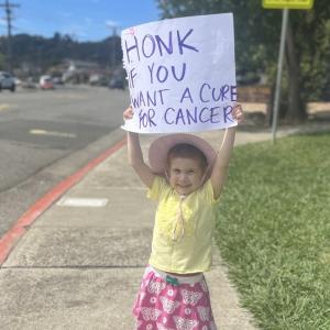 little white girl in a yellow shirt and pink skit with butterflies on it wearing a hat and holding up a sign that says honk if you want a cure for cancer