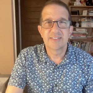 middle aged white man wearing glasses and a blue print shirt sitting in front of a bookcase