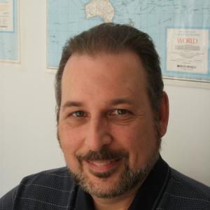 middle aged white man with a beard and mustache wearing a navy blue shirt sitting in front of a world map