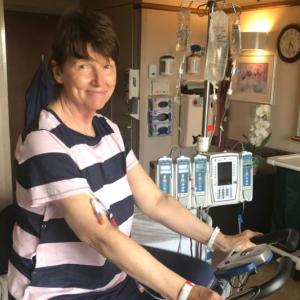 middle aged white woman with short brown hair wearing a navy and pink striped blouse and blue pants riding a stationary bike in a hospital room