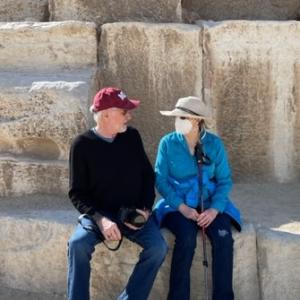 older white couple sitting in front of stones man wearing burgundy ball cap and black shirt and woman wearing blue shirt face maske and straw hat
