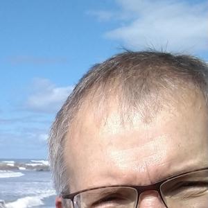 older white man with brown framed glasses plaid shirt and black jacket and big smile standing in front of the ocean