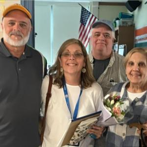 two older men wearing ball caps with a woman wearing glasses and dangly earrings holding a bouquet of flowers and their mom on the right and an American flag in the background