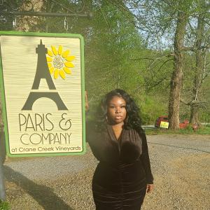 young black woman wearing a black dress with gold hoop earrings and necklace standing by a sign that says Paris and Company