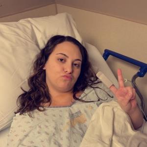 young woman with long brown hair wearing hosptial gown puckering her mouth and giving a peace sign lying in a hospital bed