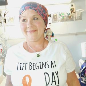 middle aged white woman wearing a colorful headscarf and white t-shirt with an orange ribbon on it sitting on a hospital bed