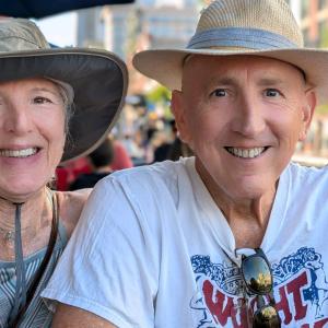 older white couple both wearing hats and smiling man wears white t-shirt with sunglasses hanging from the neck